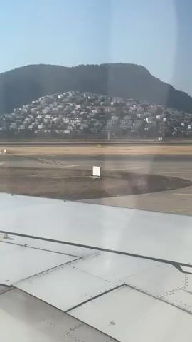 The video shows a view from an airplane window as it taxis on a runway. The plane's wing is visible in the foreground, with its reflective surface catching the sunlight. In the background, a town with white buildings is built on a hillside, with mountains behind it. The runway is visible in the middle ground, with yellow markings on the tarmac. The sky is clear and blue. The plane appears to be preparing for takeoff or has just landed.