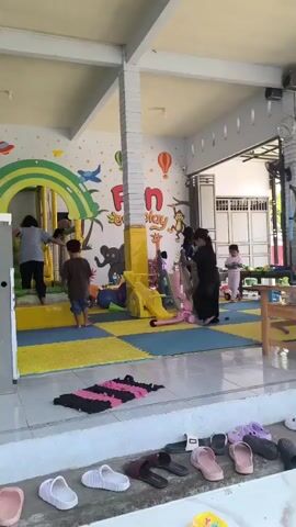 The video shows a bright indoor play area for children. The walls are decorated with colorful murals of animals, hot air balloons, and the words "Fun to Play." A large, rainbow-colored climbing structure with a slide is visible on the left. Several children are playing, including one on a scooter, another climbing on the structure, and others interacting in the play space. Adults are also present, supervising the children. The floor is covered with yellow and blue foam mats. In the foreground, several pairs of sandals and flip-flops are arranged on a ledge. The overall environment appears to be a well-equipped and safe space for children's recreation.