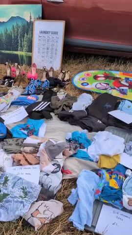 A yard sale is underway outdoors, with various items spread out on blankets and on the ground. In the background, cars are parked, suggesting a community or flea market setting. The foreground displays an assortment of goods, including clothing, children's toys, a laundry guide poster, and decorative artwork. The ground is covered in dry grass. People are visible in the background, engaged in activities related to the sale. The overall scene suggests a typical outdoor marketplace event.