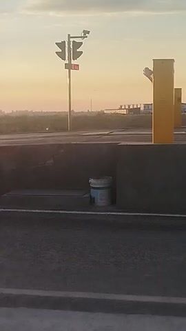 The video captures a sunrise over a landscape with some infrastructure. In the foreground, a road or paved surface is visible, with a low wall or barrier running along its edge. A white bucket with a blue lid is placed near the wall. In the middle ground, a traffic signal with multiple lights and what appears to be a camera is mounted on a pole. Further back, more poles with cameras and signs are visible. The background consists of a hazy horizon with distant buildings and terrain, under a sky with soft clouds illuminated by the rising sun. The overall impression is of an outdoor scene, possibly near a roadway or an industrial area, at dawn.