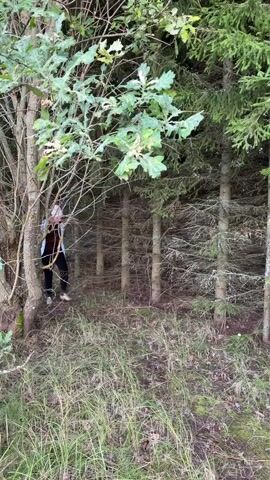 A woman is walking in a forest while holding two baskets. She is wearing a light blue jacket, a brown sweater, black pants, and a headscarf. The forest is dense with trees and undergrowth. She is walking from right to left in the frame.
