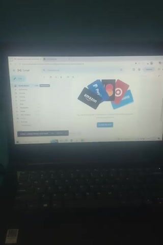 The video shows a laptop screen displaying a collection of gift cards arranged in a cluster on a white background. The gift cards include Amazon, PayPal, Mastercard, and Target. The laptop is open, and part of its black keyboard is visible in the foreground. The video is shot from a slightly overhead angle, and the lighting is somewhat dim. The scene is static, with no discernible action or movement other than the slight shift in camera perspective between frames. The environment appears to be indoors, likely on a desk or table.