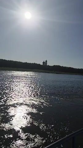 A wide shot shows a sunlit river with a distant treeline and a building with domes. The sun is high and bright, casting a glare on the water. The water is choppy, with light reflecting off the surface. A faint silhouette of trees is visible on the far bank, and on top of the trees, a building with two domes is visible. A single bird is flying in the sky. The bottom of the frame shows a railing.