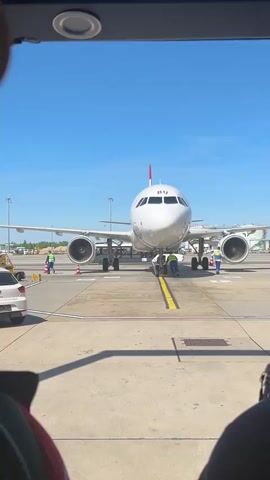 The video shows an airplane with the letters "BU" on its nose, seen through the window of a bus or similar vehicle. An airport worker in a yellow vest is kneeling in front of the plane, while other workers are nearby. The sky is clear and blue. In the foreground of the shot, a person is visible, looking down at something in their hands and wearing white earbuds. The lighting is bright and sunny.