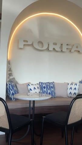 The video shows a cafe interior with a booth seat in the background. The booth has several cushions with a blue and white pattern. Above the booth, a sign reads "PORFA". A circular stone accent is visible to the left of the sign. A small round table with a marble top is in front of the booth, with two chairs on either side. The chairs have black frames and woven seats and backs. A neon light illuminates the archway above the sign. The lighting suggests it is daytime.