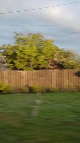 The video shows a blurry, side-view, exterior shot of a residential area with houses and trees. The camera appears to be moving, possibly from a vehicle, causing the motion blur. The environment appears to be rural or suburban. There are no discernible people or specific actions occurring.