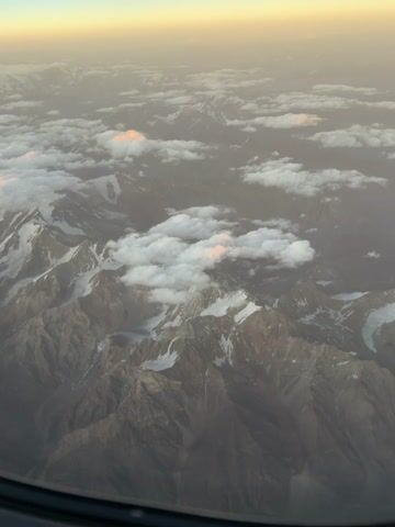 The video shows an aerial view of a mountain range with snow-capped peaks and clouds. The sun is setting, casting a warm glow on the clouds and the horizon. The camera appears to be inside an airplane, as evidenced by the curved window frame at the bottom of the screen. The mountains are rugged and appear to be covered in dry, rocky terrain with patches of snow and ice. Some clouds are scattered among the peaks, with a few appearing to be lit by the setting sun. The sky transitions from a pale yellow near the horizon to a light blue.