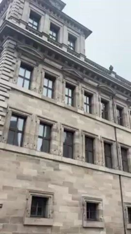 The video shows a low-angle view of a multi-story stone building with numerous windows under an overcast sky. The building features intricate architectural details, including decorative moldings and a central tower with a green spire. The camera slowly pans upward, revealing more of the building's facade. The overall impression is one of historic European architecture on a cloudy day.