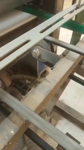 The video shows a close-up view of a conveyor belt system. A white material, possibly paper or plastic sheeting, moves along the belt. The belt is driven by a pulley system with black belts and metal components. The surrounding environment appears to be an industrial setting with metal framing and some exposed wiring. The footage is shaky, suggesting it was filmed while the machine was in operation.