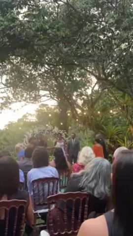The video shows a wedding ceremony taking place outdoors, with guests seated on wooden chairs facing the front. A bride in a white dress and a groom in a suit stand at the altar, accompanied by several people, possibly the wedding party. The setting is a garden with lush green trees and foliage, with string lights hanging from the trees in the background. The scene appears to be filmed from the perspective of a guest. The wedding party bows their heads in a solemn gesture.