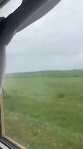 The video captures a view through a window, likely from a moving vehicle such as a train or bus. The window frame is visible in the foreground, with a white object, possibly a sunshade or part of the vehicle's interior, partially obscuring the upper left corner. Outside the window, a vast expanse of green fields stretches towards the horizon under a cloudy, overcast sky. The landscape appears rural and open, with sparse vegetation and faint signs of human infrastructure like utility poles in the distance. The footage is shot from a slightly low angle, emphasizing the expansive view of the fields. The lighting suggests it is daytime, but the overcast conditions create a muted, diffused light. There are no people or distinct actions visible, and the focus is on the passing scenery.