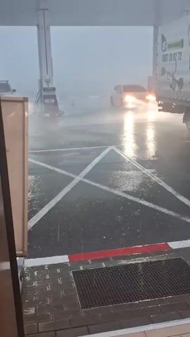The video shows a gas station forecourt experiencing extremely heavy rain, causing significant pooling of water on the ground and reducing visibility. A white sedan drives slowly through the downpour, its headlights reflecting brightly on the wet surface. To the right, a white truck is visible, featuring a "GARDEN" logo, the phone number "0887 29 62 72", and images of gardening equipment like a lawnmower and a tiller on its side. Gas pumps are visible under a protective canopy to the left, with some numbers like "5" and "15" partially discernible on one. The scene depicts severe weather conditions impacting the area, with everything appearing hazy due to the intense precipitation.