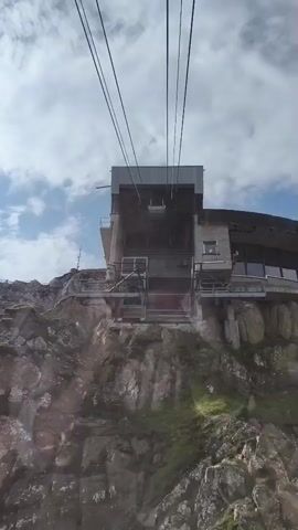 A view from inside a cable car looking up at a cable car station perched on a rocky mountainside. The station is a modern, multi-level structure with large windows and metal balconies. Several thick cables run overhead, presumably supporting the cable car system. The sky is cloudy with patches of blue. The rock face below the station is rugged and covered in sparse vegetation. The cable car appears to be ascending.