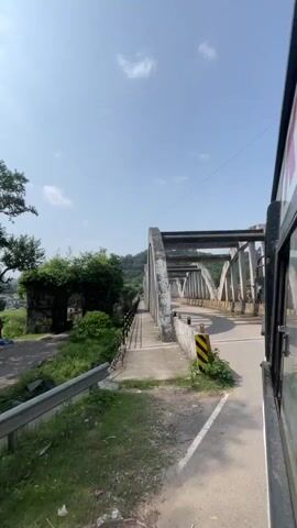 A car is driving over a bridge. The bridge is made of concrete and steel and has a sidewalk on the left side. There are trees and hills in the background. The sky is blue and there are some clouds.