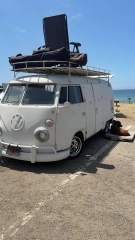 The video shows a white Volkswagen van parked on a gravel lot near a beach. A dog is lying on a cushioned mat next to the van. The dog appears to be a dark brown color with some lighter markings. The van has a roof rack with items on it, including a large black chair and some bags. The ocean is visible in the background, with a few people walking on the beach and some picnic tables. The sky is blue and clear, indicating a sunny day. The camera pans slightly to the right, focusing on the dog and the side of the van.