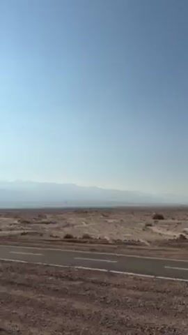 The video shows a desert landscape under a clear, light blue sky. In the distance, a hazy mountain range stretches across the horizon. A paved road with white lines runs across the foreground of the scene. The land is barren and rocky, with sparse vegetation visible. The camera appears to be in motion, likely from a passing vehicle on the road.