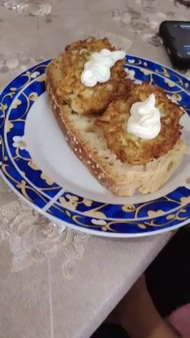 The video displays a close-up shot of a slice of bread topped with two fritters and dollops of mayonnaise. The bread appears to be a rustic loaf, possibly sourdough, with sesame seeds on its crust. The fritters are golden brown and seem to be made from grated vegetables, possibly zucchini or potatoes. Two distinct swirls of mayonnaise are placed on top of the fritters. The plate has a blue and white patterned rim. In the background, a table surface with a textured tablecloth is visible, along with a corner of a smartphone. The lighting is soft, suggesting an indoor setting.
