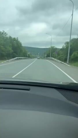 The video shows a view from inside a car, looking out the windshield. The car is driving on a two-lane highway with trees and rolling hills in the background. The sky is cloudy. There are guardrails on both sides of the road. There are streetlights along the side of the road. A sign with Russian text "РАЗДОЛЬНОЕ" is visible. Another car is in the distance, also driving on the highway. A fuzzy dark object with eyes is visible on the dashboard in the foreground. The video lasts for about five seconds.