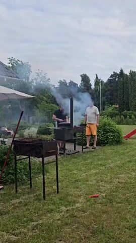 The video shows a group of people enjoying a barbecue in a backyard setting in Harkujärve, Estonia. Two men are actively grilling, with one tending to the barbecue and another, wearing an orange t-shirt, standing nearby. Smoke billows from the grill, indicating that food is being cooked. In the background, other individuals are gathered around a table under a large umbrella, suggesting a social gathering. The scene appears to be set in the afternoon, with a cloudy sky overhead. Lush green grass covers the ground, and various plants and trees surround the area, including a pine forest visible in the distance. Two brooms are propped up near the barbecues, and a yellow leaf-sweeping tool is leaning against a low wall. The atmosphere seems relaxed and casual, typical of a summer barbecue. There is no visible text in the video.