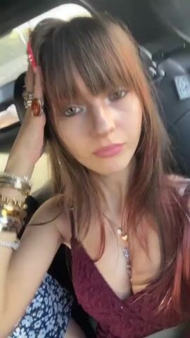 A young woman with long brown hair and bangs, wearing a burgundy dress and several rings on her fingers, is in the passenger seat of a vehicle. She is holding up her hand to show off her long, colorful, pointed fingernails. She appears to be moving her mouth as if speaking or singing. In the background, a bottle of drink is visible, and another person, partially obscured, is in the seat next to her. The lighting suggests it is daytime.