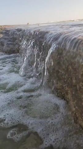A close-up shot focuses on a shallow waterfall cascading over a rocky edge into foamy water below. The water is clear, and the sunlight reflects off its surface, creating a shimmering effect. The rocks appear rough and textured, with some small shells or debris visible. The background is out of focus, showing a hazy expanse of water and a clear sky. The scene suggests a natural outdoor environment, possibly a coastline or a stream. The video is very brief, lasting only a few seconds, and shows the water flowing consistently over the edge. There are no people or animals visible in the frame.