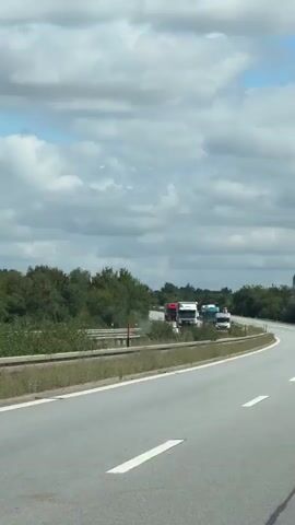 A video clip shows several semi-trailer trucks stopped on a highway. The trucks are in lanes and appear to be stationary. The surrounding environment consists of trees, a grassy median, and a cloudy sky. The camera is moving, indicating it is likely from another vehicle on the highway. The trucks are varied in color and make, including a white MAN truck with red accents, a white Iveco truck, a blue and white DAF truck, and others. The scene suggests a traffic jam or a standstill on the road.