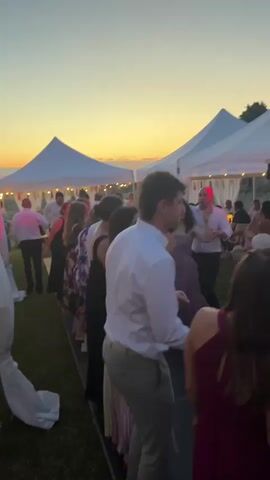 A video depicts an outdoor evening event under white tents with string lights. A diverse group of people, dressed in semi-formal attire, dance in a line on a dark platform. The sky in the background displays warm sunset colors. The camera moves slightly, showing more of the setup, including white fabric draping poles and green grass.