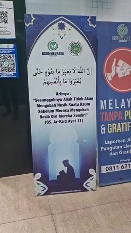 A vertical, rectangular banner with Islamic calligraphy and artwork is displayed in what appears to be a public indoor space, possibly a hospital or government building, given the presence of other signage with QR codes and official logos. The banner itself features a large, arched design at the top with floral motifs and the logos of "RSUD MEURAXA" and another green circular logo with Arabic text. Below the logos, there is Arabic script, followed by its Indonesian translation in quotes. The translation reads, "Indeed, Allah will not change the condition of a people until they change themselves." This quote is attributed to "QS. Ar-Ra'd Ayat 11." At the bottom of the banner, a silhouette of a person is depicted praying or reading within an arched doorway. The colors used are primarily blue, white, and some purple hues, creating a serene atmosphere. To the right of this banner, a portion of another blue banner is visible, which seems to advertise services from "RSUD MEURAXA" and includes text about reporting illegal levies and a phone number. The floor appears to be tiled. The lighting is moderate, with some shadows cast on the floor. The overall scene suggests an emphasis on faith and self-improvement within an institutional setting.