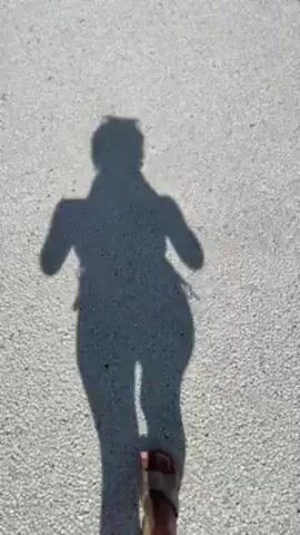 The video begins with a person walking along a white, speckled concrete path, their shadow visible on the ground. As the camera moves forward, the scene widens to reveal a beautifully landscaped area. Two modern, wooden-clad buildings with black roofs are visible on either side of the path. A body of water stretches out behind the buildings, reflecting the blue sky with scattered clouds. Lush green grass surrounds the path and buildings, interspersed with various plants and trees. The overall atmosphere is peaceful and serene.