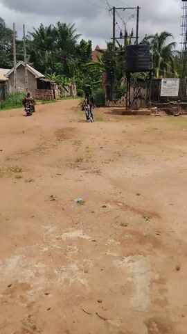 The video shows a wide, unpaved dirt road under an overcast sky. Two motorcycles are visible on the road. One motorcycle, with a single rider, approaches the camera from a distance. Another motorcycle is further down the road, moving in the same direction. On the left side of the road, there are simple, low-rise buildings and lush green vegetation. On the right, there is a structure with a large black water tank elevated on a metal frame, next to a gate and a sign (text illegible). Utility poles and power lines are also visible on the right. The scene depicts a rural or semi-urban setting, likely in Akwa Etiti, Nigeria, during the daytime.