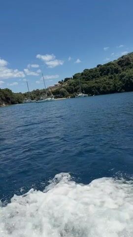 A video captures a view from a moving boat on a bright, sunny day. The water is a deep blue, with white foam trailing from the boat's wake in the foreground. In the middle ground, several sailboats are anchored in a calm bay. The shoreline is covered in lush green trees and vegetation, with some buildings visible on the hillsides. The sky is a clear blue with a few scattered white clouds. The camera is angled slightly upwards, emphasizing the vastness of the sky and the expansive view of the bay. The video appears to be shot during daylight hours.