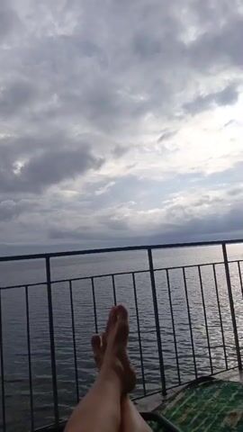 The video shows a view from a balcony or deck overlooking a body of water. The camera is angled upwards, focusing on the sky which is filled with dramatic, grey clouds. Sunlight is breaking through the clouds in several places, creating bright patches and reflecting on the water. In the foreground, a railing separates the viewer from the water. Below the railing, a person's bare feet and legs are visible, crossed at the ankles. The person appears to be lying down or reclining, with a green and white striped blanket visible beneath them. The overall atmosphere is one of relaxation and enjoying the view of the overcast sky and water.