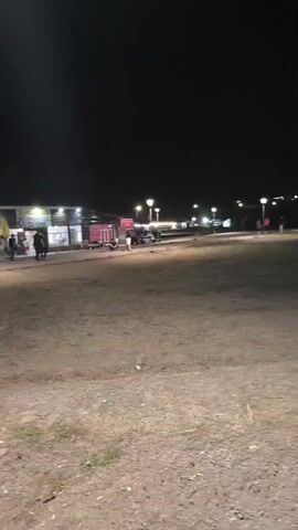 The video shows a night scene with a dark sky. In the background, there are several illuminated structures that appear to be businesses or shops. There are also streetlights visible. In the foreground, there is a patch of dry grass and dirt. Several people are present in the background, walking and standing near the illuminated areas. There are also some vehicles visible. The overall impression is that of an outdoor area with businesses operating at night.