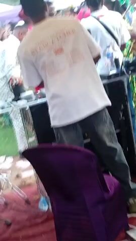The video shows a live band performing under a white tent, with a crowd of people gathered around. The band includes a singer in a red outfit, a saxophone player, and a DJ. The crowd includes people of various ages and genders, dressed in colorful clothing. Many of them are recording the performance on their phones. The environment appears to be an outdoor event, possibly a wedding or a festival. There is green grass visible, and the tent provides shade for the performers and the audience.