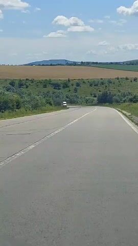 A white van drives on a paved road through a rural landscape under a partly cloudy sky. The road curves to the left. Fields of dry grass and green bushes are visible on either side of the road, with rolling hills in the background.