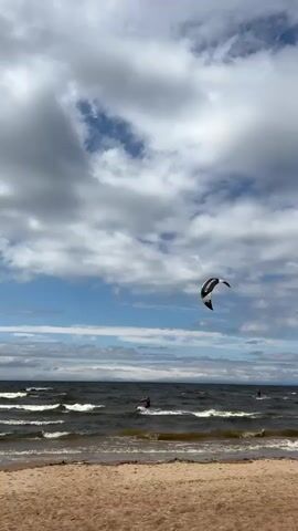 The video shows a view of a beach and the ocean. The sky is partly cloudy with patches of blue sky. In the distance, there are several people kite surfing on the water. In the foreground, a sandy beach is visible. The waves are choppy and breaking on the shore. The camera is positioned on the beach, looking out towards the ocean.