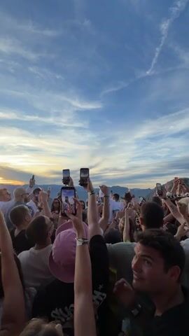 A group of people stand outdoors, most with their arms raised, holding up smartphones as if to record or photograph something above them. The sky is a gradient of blues and oranges, with clouds streaking across it. Mountains are visible in the distance. In the center of the frame, a few individuals are standing closer together, appearing to be the focus of attention. One person, wearing a white cap and a white shirt with blue trim, is central. A drone is visible in the upper portion of the frame, appearing to be in flight. The overall atmosphere seems to be one of excitement or observation.
