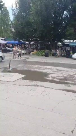 A paved outdoor market scene is depicted. A silver hatchback car is parked on the left side of the frame. Two people, an adult and a child, are walking across a puddle-filled area in the center of the frame. In the background, several market stalls with blue and white canopies are visible, along with trees and more people browsing. The ground is cracked and uneven.
