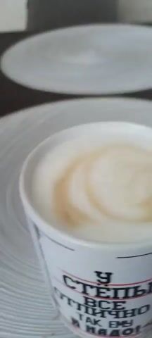 A close-up shot shows a white mug filled with foamy coffee. A hand holding a spoon is seen stirring the coffee, creating swirls in the foam. The mug is placed on a white plate with a textured, circular design, which in turn is on a darker surface. Another similar plate is visible in the background, out of focus. The text on the mug is partially visible, with some Cyrillic letters. The lighting is bright, suggesting an indoor setting.