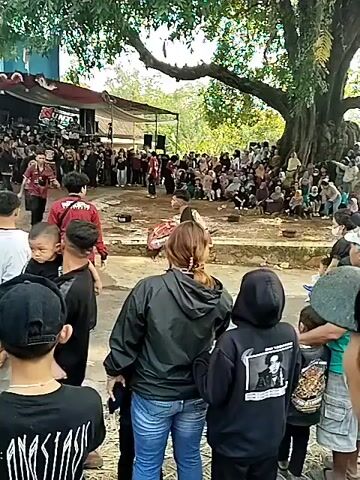 A large crowd gathers outdoors under a massive tree, watching a performance. In the foreground, several people face away from the camera, looking towards the activity. To the left, a stage with sound equipment is set up, and a group of people are visible on it. In the center of the scene, a performer in a colorful costume is dancing. The ground appears to be dirt. The atmosphere is lively, with many spectators present.