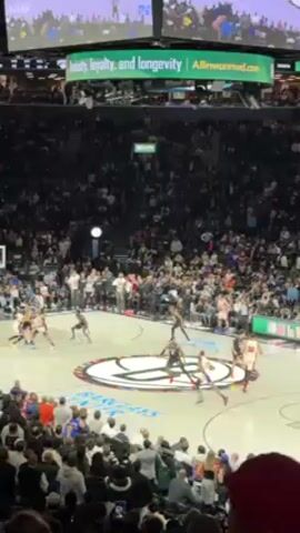 Here is a description of the video:

The video shows a basketball game in progress at an indoor stadium. Players in black and white uniforms and red and white uniforms are on the court. The stadium is packed with spectators. A large screen displays ads and replays during the game. The court has a logo in the center. The camera captures the scene from an elevated spectator perspective. The environment is well-lit with stadium lighting.