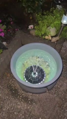 The video shows a garden at night. In the foreground, there is a shallow blue basin filled with green water and a solar-powered fountain. The fountain has a circular base with lights that illuminate the water with a green glow. The fountain is spraying water upwards in a circular pattern. The basin is surrounded by rocks and some greenery. In the background, there are various solar-powered garden lights, including lights shaped like flowers and mushrooms, as well as string lights. There are also pink flowers and green bushes. The overall scene is one of a decorated garden illuminated by artificial lights at night.
