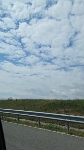 The video shows a view from a moving vehicle on a highway. A silver guardrail runs parallel to the road. Beyond the guardrail, there is a grassy slope dotted with vegetation. The sky is predominantly blue, filled with numerous white, fluffy clouds. The camera appears to be positioned low in the car. Electronic dance music plays.