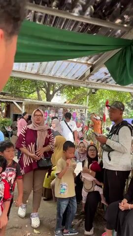 A group of people are gathered outdoors under a makeshift canopy. The canopy appears to be made of a green fabric stretched over a metal frame. Sunlight filters through the leaves of nearby trees. In the foreground, a person's back is to the camera. To the left, a woman wearing a maroon and white striped shirt and a light pink hijab is looking to her right. Next to her, a young boy in a tan shirt and blue jeans is looking down. To the right of the boy, a woman in a black hijab is holding a baby wearing a pink outfit. A man in a white shirt and a dark backpack is standing behind them. Further back, other people are visible, including a man in a blue denim jacket and a woman in a blue hijab. There are stalls or vendors in the background, with colorful items displayed. The overall impression is of a casual gathering or a small market.