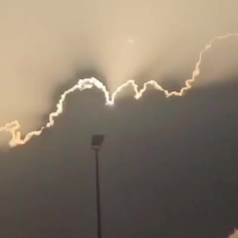 A video shows a streetlamp silhouetted against a cloudy sky at sunset or sunrise. Rays of sunlight are breaking through the clouds, creating a dramatic effect. In the background, a building with signage is visible. The camera pans slightly, capturing different angles of the sky and the surrounding environment.