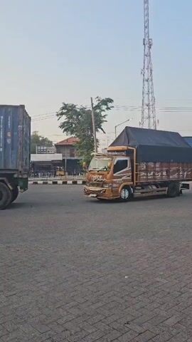 A long shot shows a large blue shipping container on a trailer with a red semi-truck attached, parked under a canopy. The trailer has several axles and wheels. A smaller orange truck with a black tarp over its load is visible in the background, turning right on a street. The ground is paved with interlocking bricks. A tall radio tower is visible in the distance against a hazy sky. Trees and buildings are also in the background. The time stamp at the bottom of the screen indicates the video is 5 seconds long.