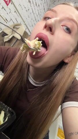 A blonde woman eats pasta from a black plastic container. She uses a fork and looks up at the camera while she eats, and after taking a bite, closes her eyes briefly and appears to savor it. She then sticks her tongue out. She is wearing a brown shirt, a gold necklace, and has a tattoo on her left forearm. The background includes a white brick wall, a coat hanging on a rack, and a sign.