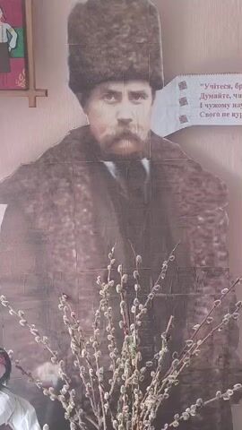 A life-sized cardboard cutout of Taras Shevchenko, a Ukrainian poet and artist, stands in front of a light-colored wall. He is depicted wearing a dark fur hat and a dark coat, with a mustache and beard. To the right of the cutout, a strip of paper with text in Ukrainian is attached to the wall. In front of the cutout, a bouquet of pussy willow branches with catkins is arranged.