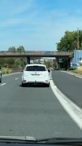 This is a vertical video taken from inside a vehicle traveling on a highway. The primary focus is on a white Ford Focus in front, also traveling on the highway. The Ford Focus has two people with their arms extended out of the open windows on opposite sides of the vehicle. The person on the left has their arm extended with their hand reaching out. The person on the right has their arm extended and is waving. The license plate of the Ford Focus is visible as BA 624 OG. In the background, there is an overpass with graffiti, trees, and buildings. Another white car is visible in front of the Ford Focus. The sky is blue and it appears to be a sunny day. The highway has multiple lanes of traffic.