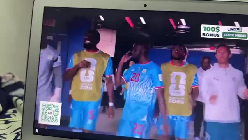 A laptop screen displays a video of three male athletes walking in single file down a brightly lit corridor. The athlete in the foreground wears a blue jersey with the number 20 on it. To his left, the second athlete wears a yellow vest over a blue jersey and the third athlete, on the far left, is a goalkeeper in a grey jersey. A graphic overlay at the bottom left of the screen displays "VOANE WISSA 9th GOAL FOR CONGO DR." The walls of the corridor are decorated with large FIFA World Cup 2026 branding. An advertisement for "EARN BIG" is visible on the upper right side of the screen.